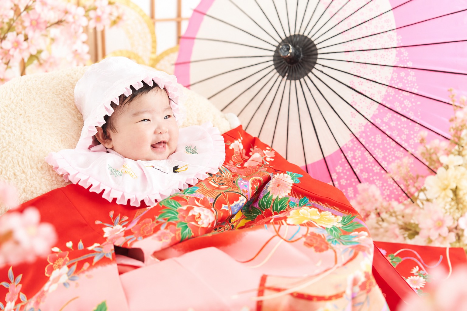 白いフードと華やかな赤い産着に包まれ、笑顔を見せる赤ちゃん